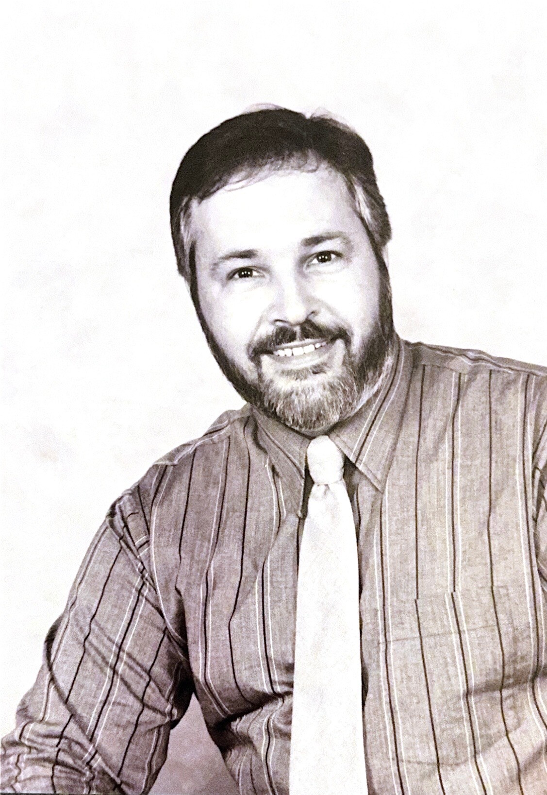 Smiling man with short dark hair and beard in a striped shirt and light tie, posing in a studio portrait (black-and-white photo).