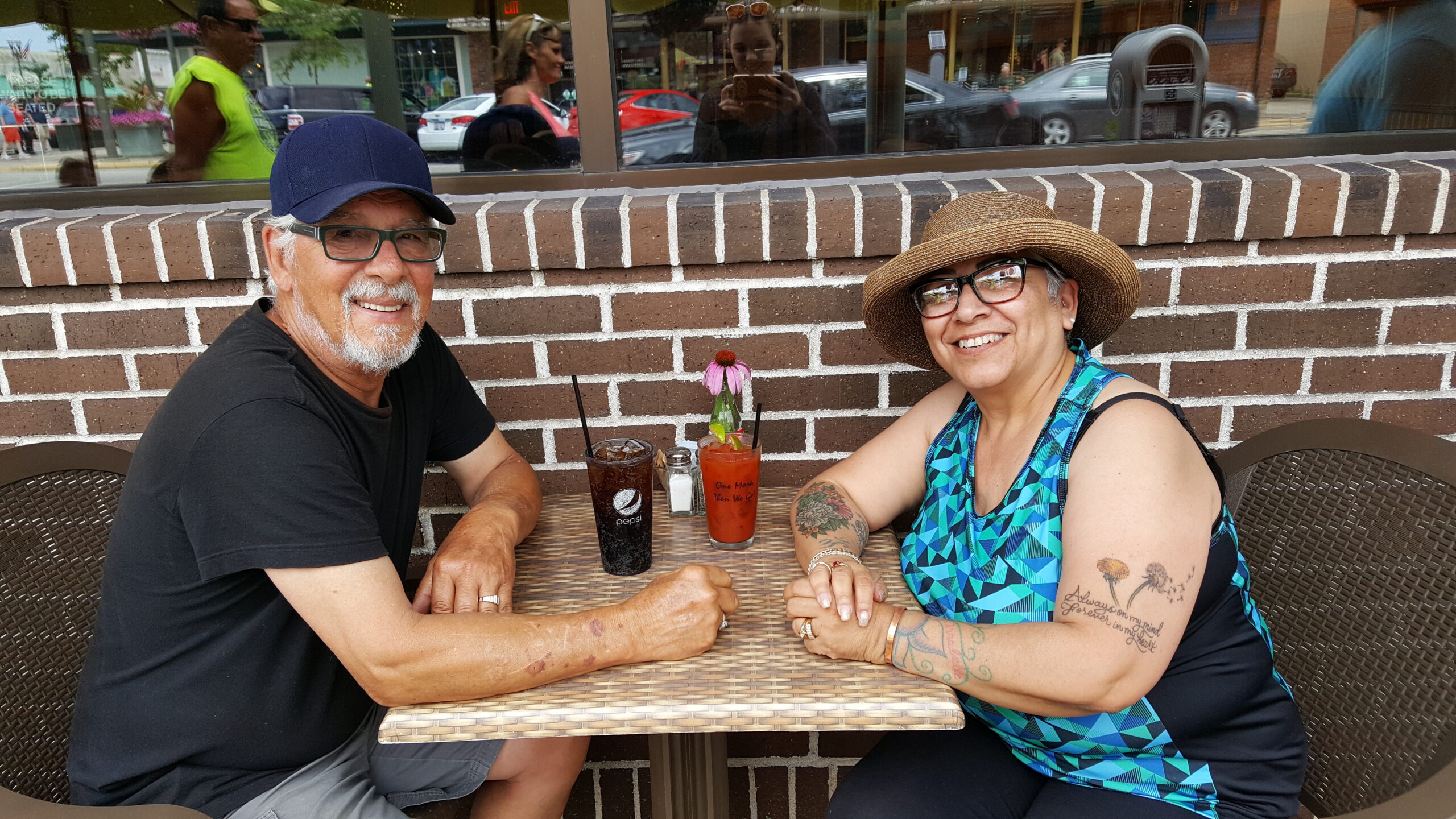Smiling man and woman sit at an outdoor cafe table, two drinks with a pink flower garnish against a brick wall behind them.