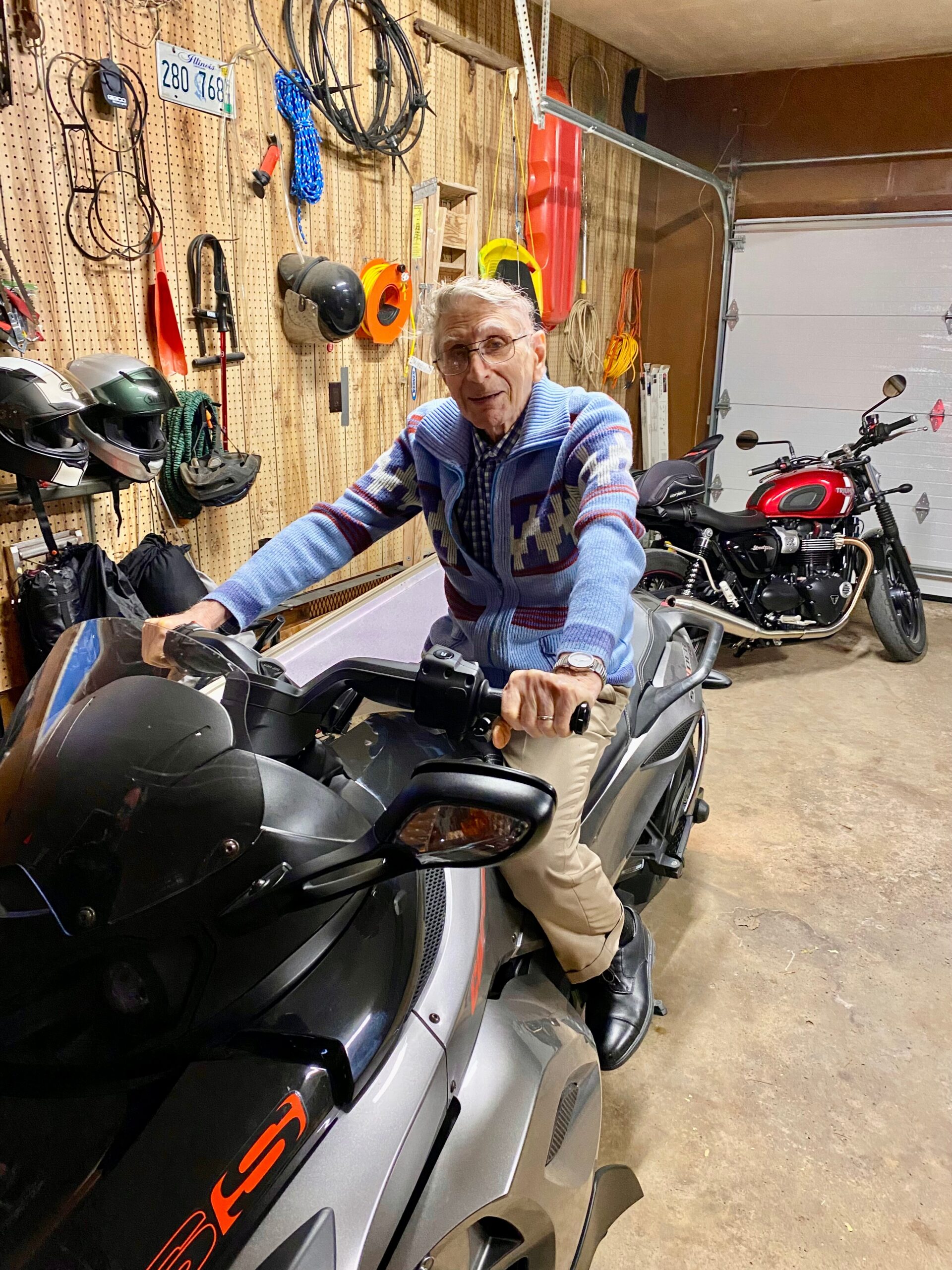 Older man wearing a blue sweater rides a gray motorcycle in a cluttered garage with tools and a red bike in the background.