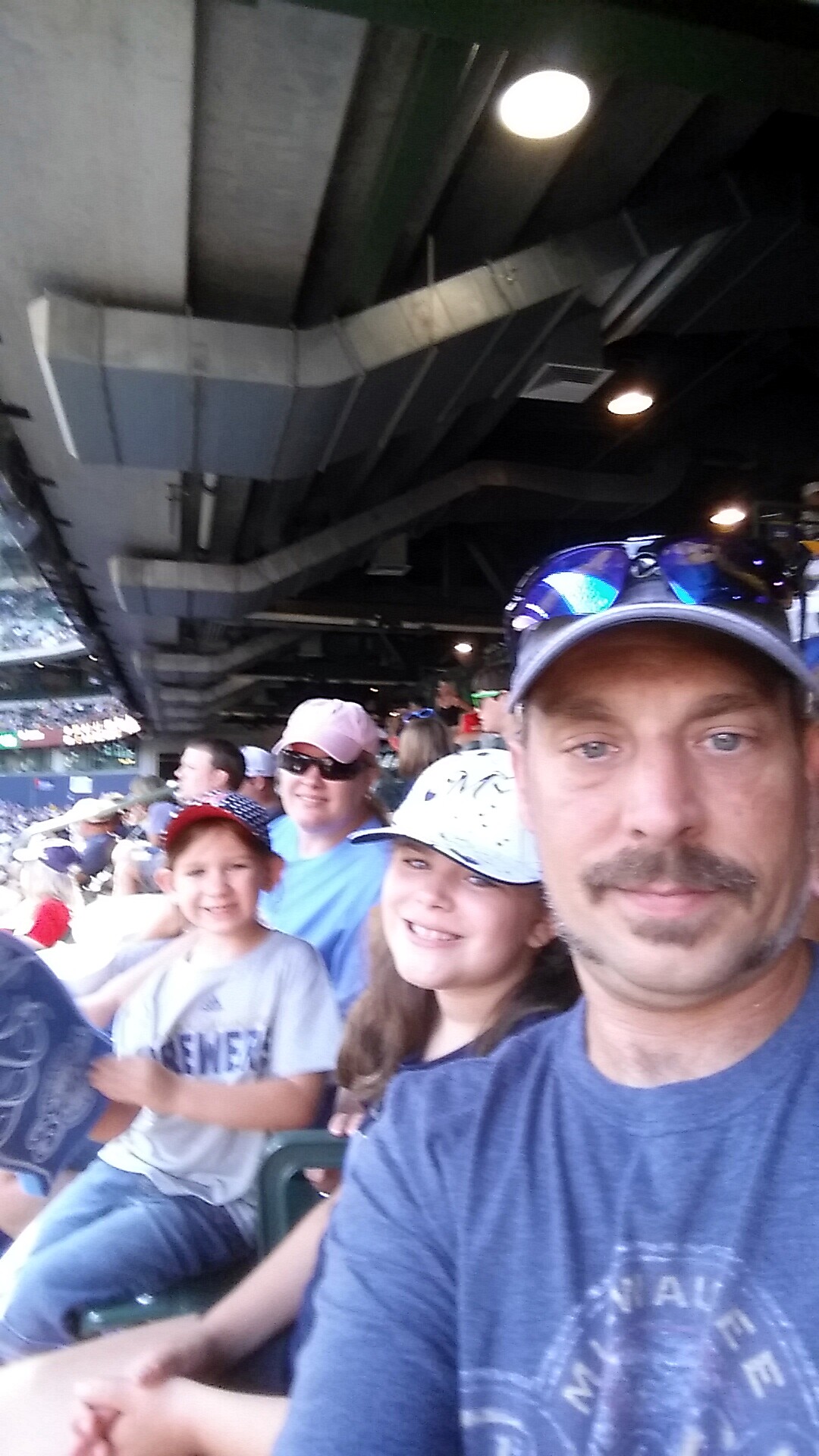 Man in a blue shirt taking a selfie at a stadium, with smiling kids and fans behind him under a dark, industrial ceiling.
