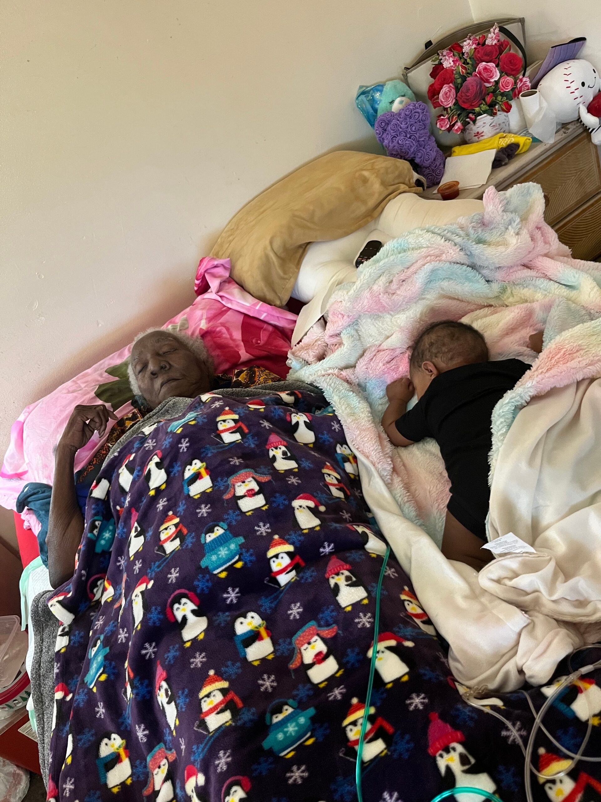 Two people asleep on a bed, elderly man on the left and a child on the right, covered with colorful blankets and a penguin-themed quilt sectioned with pink pastel throws.