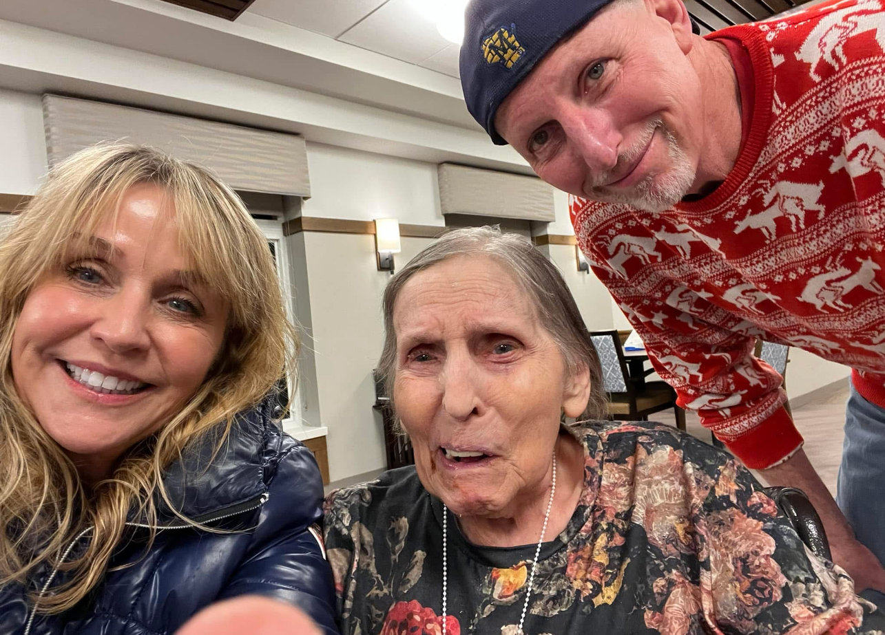 Three people posing for a selfie indoors: a smiling blonde woman on the left, an elderly woman in the center with a floral top, and a man in a red patterned sweater leaning in from the right.