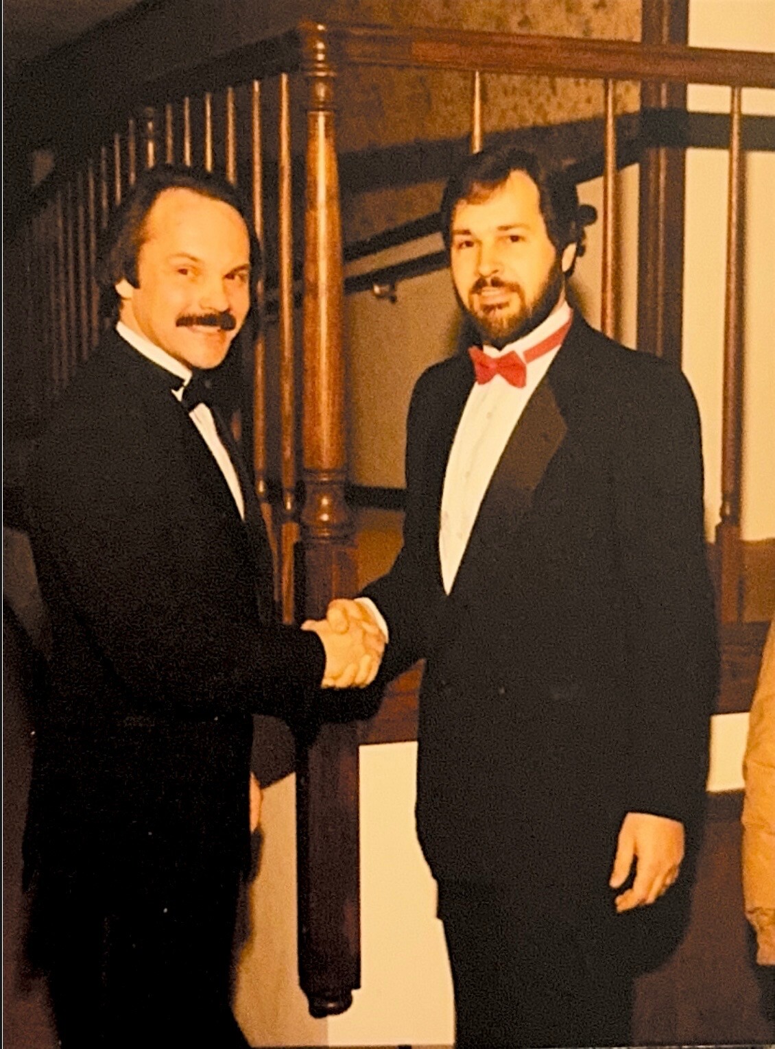 Two men in tuxedos shake hands and smile at the camera in a formal setting with a wooden railing behind them.