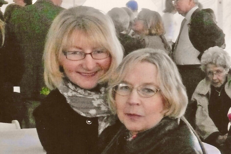 Two women wearing glasses pose for a photo at a crowded indoor gathering.