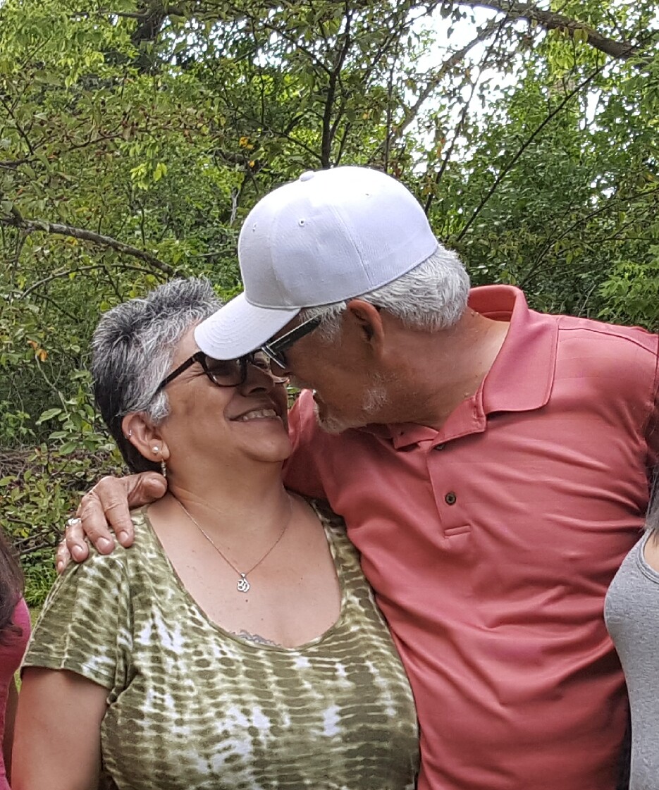 Older couple sharing a kiss outdoors, smiling with arms around each other in a green setting.
