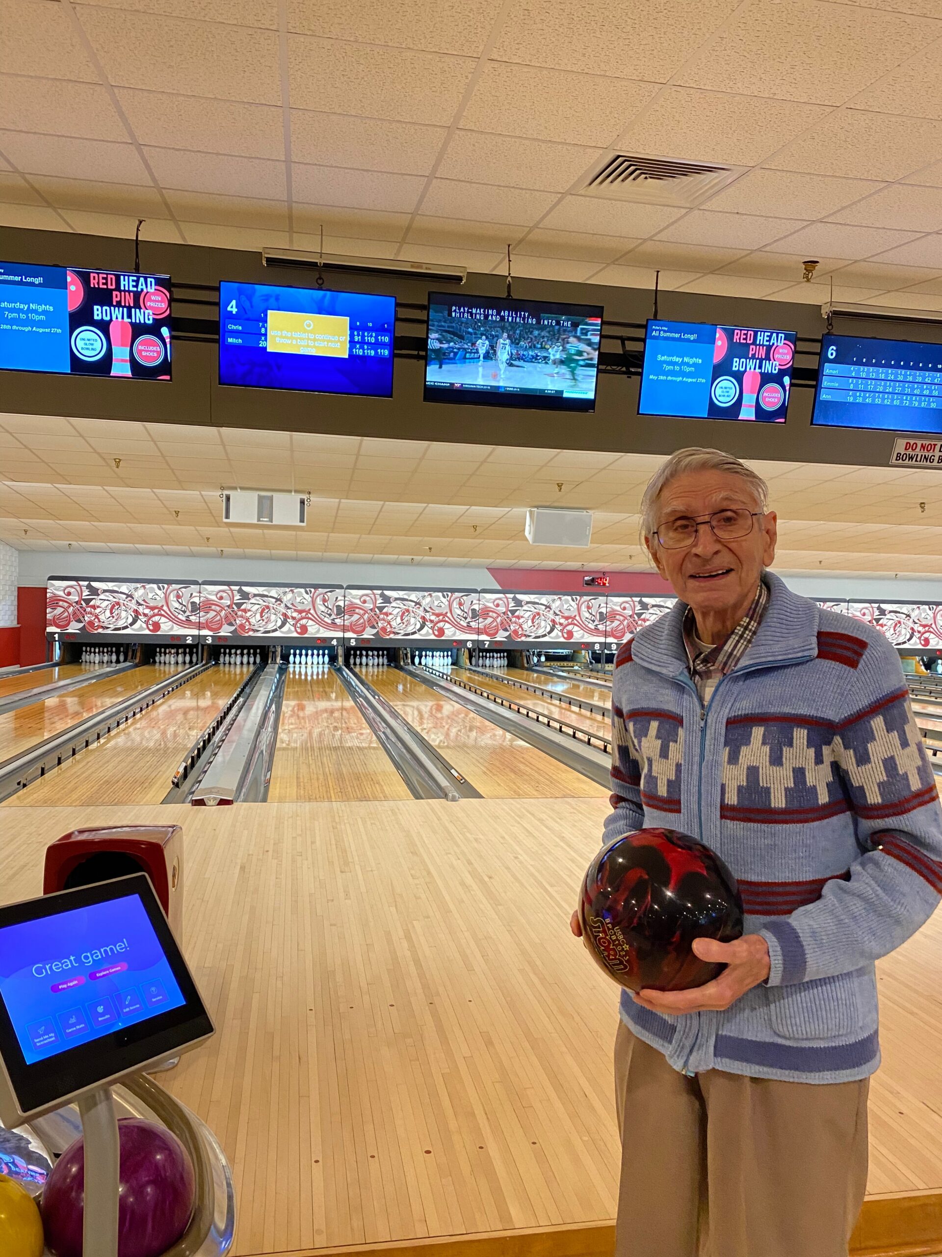 Elderly man in a blue sweater holds a red bowling ball at the bowling alley, smiling at the camera.