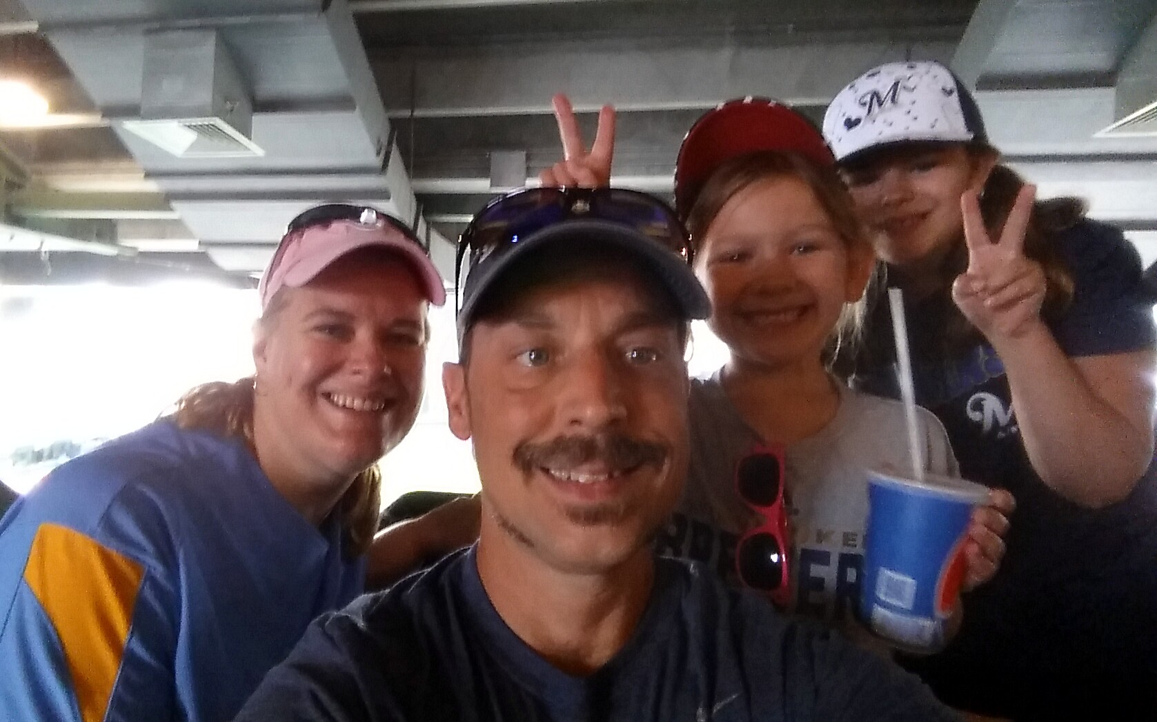 Four friends pose for a selfie indoors; man center-front, woman left in pink cap, woman right with white cap making peace sign, one holding a blue cup with straw