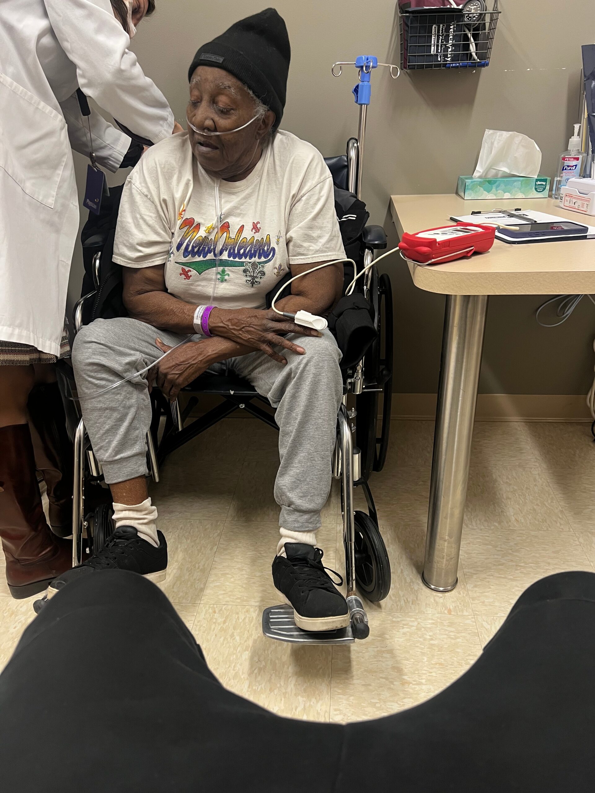 Elderly woman in a wheelchair with nasal oxygen, wearing a beanie and a colorful New Orleans shirt, in a clinic with a nurse nearby.