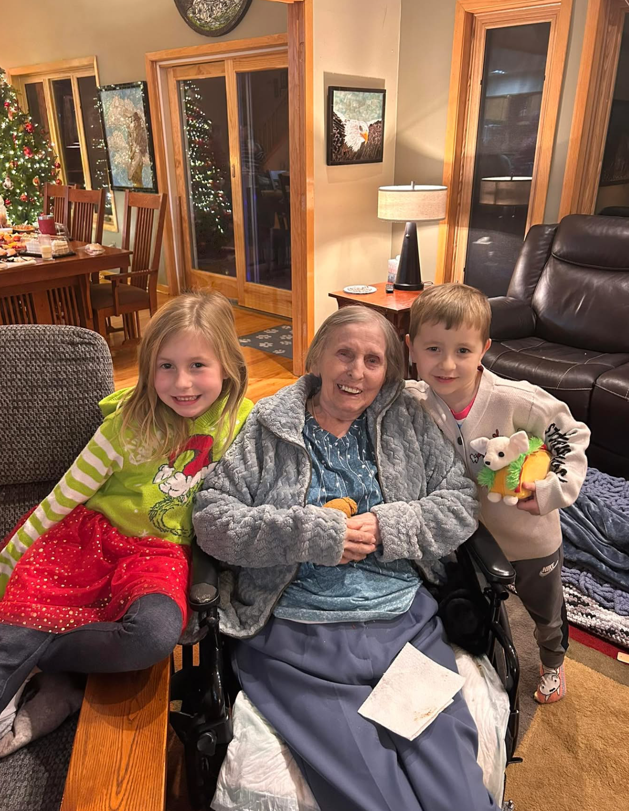 Granddaughter and grandson sit with their elderly grandmother in a wheelchair at a festive, Christmas-decorated living room; smiling together on a wooden coffee table surrounding them.
