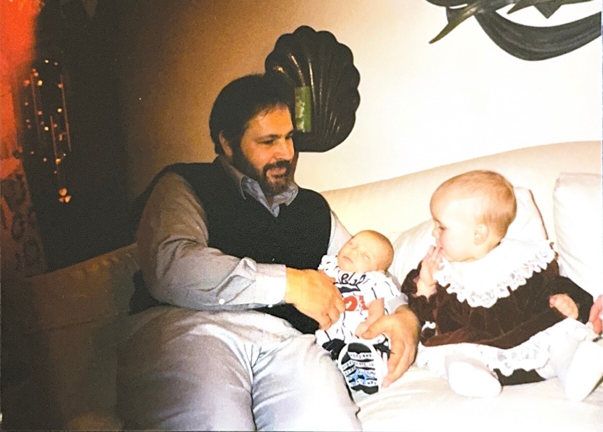 Father with a beard sits on a couch holding a sleeping baby, while a toddler girl in a brown dress with a lace collar watches nearby on the same sofa.
