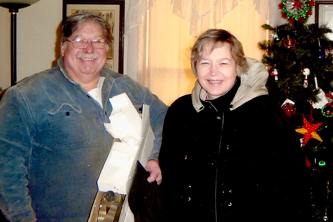 Man in a denim shirt and a woman in a black coat pose indoors, the man holding a large boxed gift near a Christmas tree.