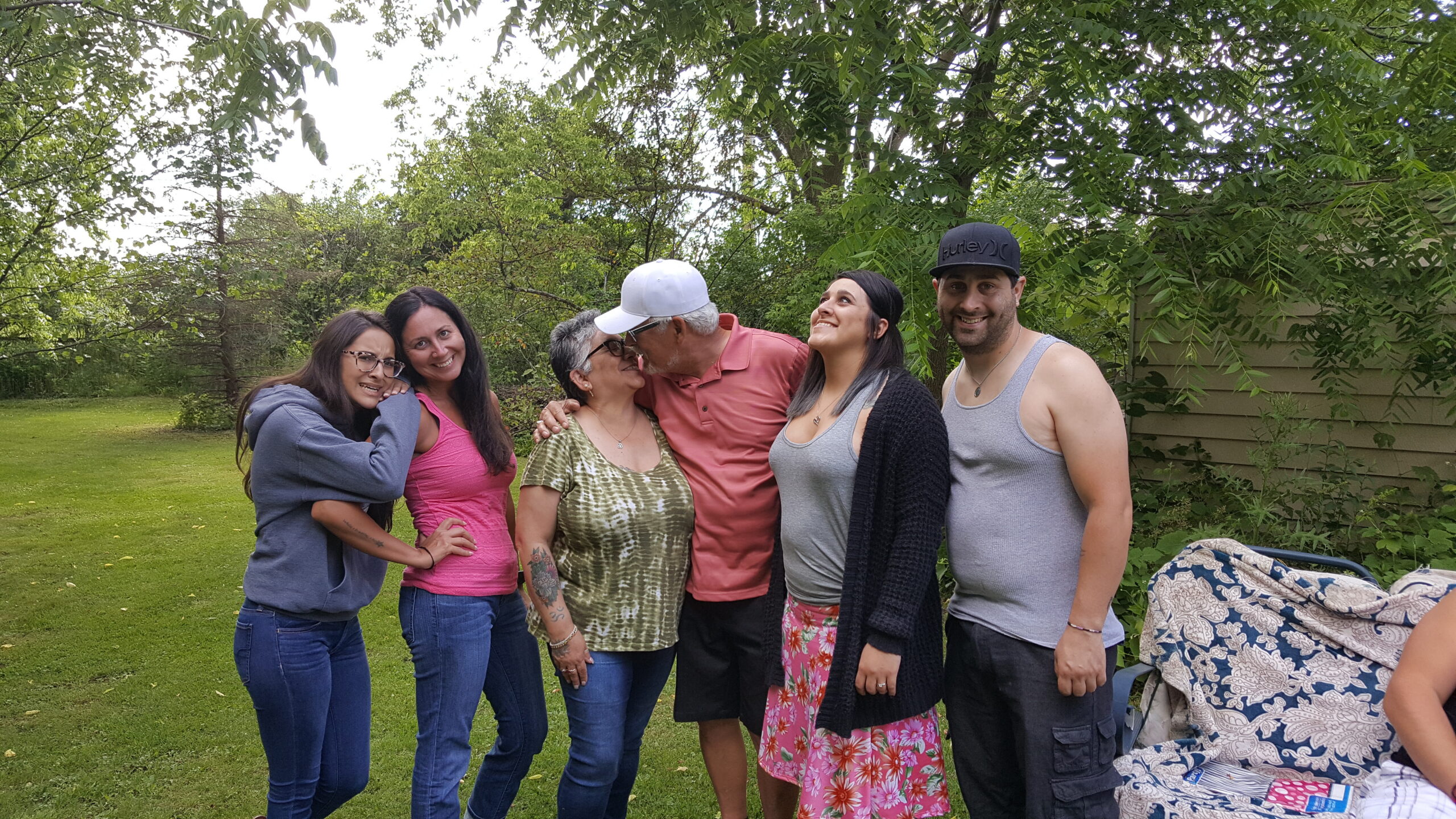 Group of six adults posing together in a backyard, smiling under leafy trees on a sunny day.