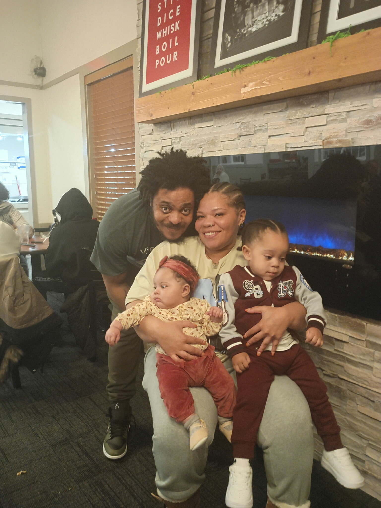 Family group photo: mother cradling an infant while a man leans in beside her and a toddler sits on her lap, in a cozy living room with a stone fireplace behind them.