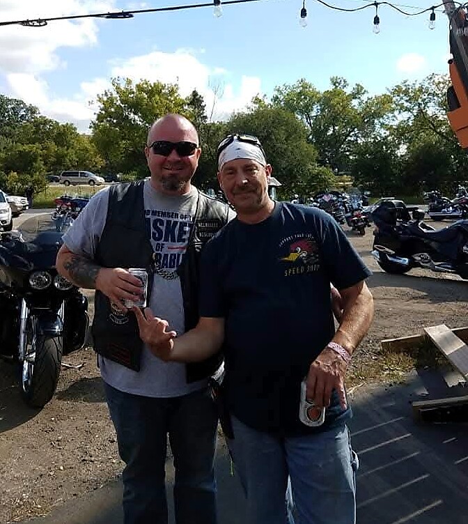 Two men posing together at a motorcycle meetup, one wearing a vest and sunglasses and the other with a bandana, both holding drinks.