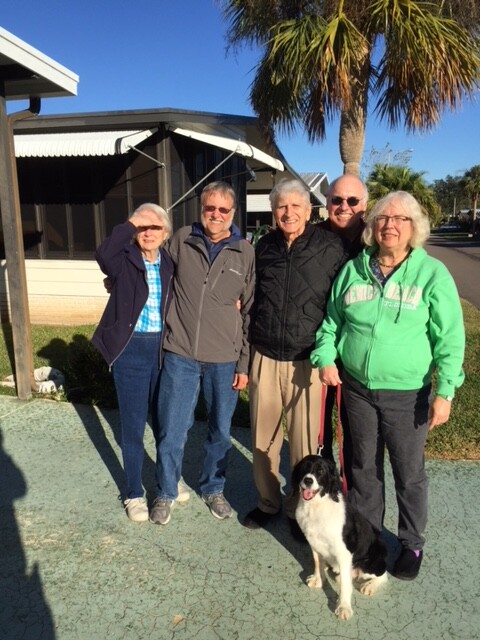 Five older adults stand together outside on a sunny day, with a black-and-white dog on a leash at their feet.