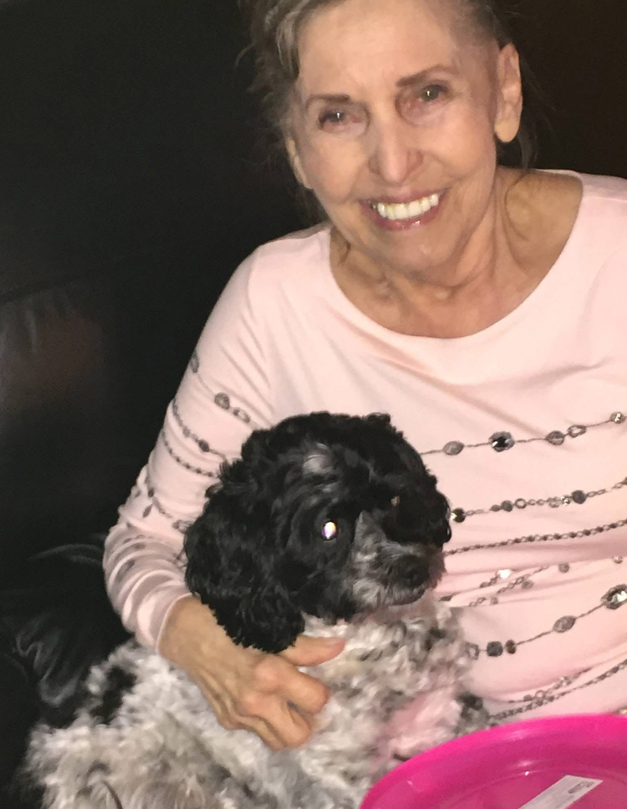 Smiling older woman in a pink top holding a fluffy black-and-white dog at night.