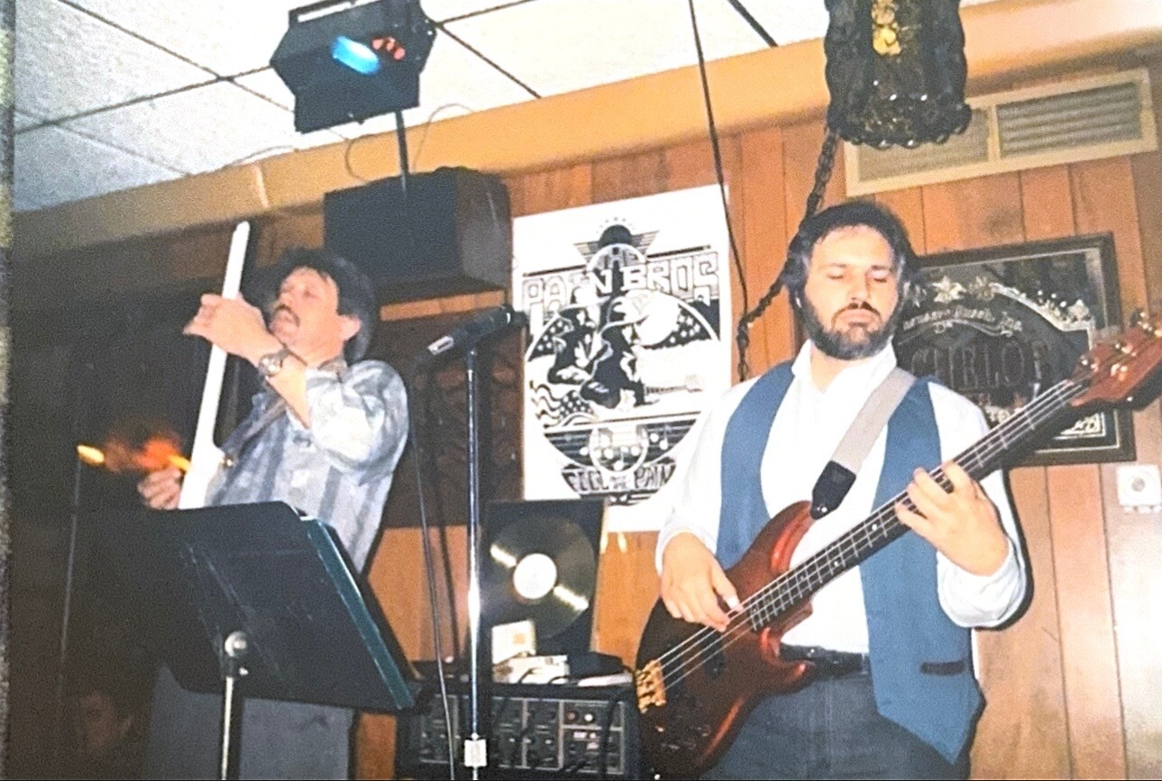 Two musicians perform on a small stage: a vocalist with a microphone and a bassist plucking a brown bass guitar.