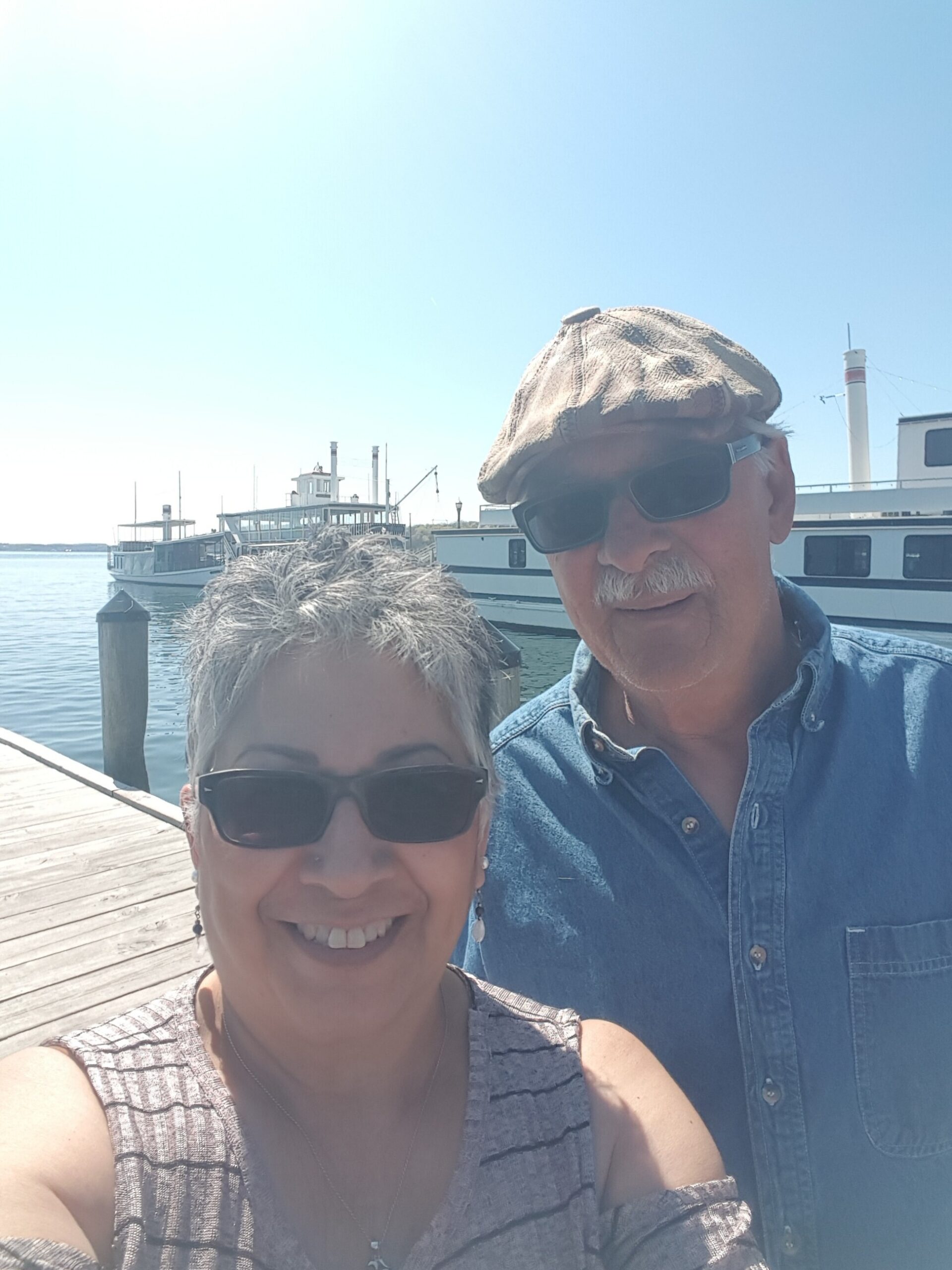 Two people wearing sunglasses take a sunny selfie at a marina with boats in the background, blue sky above.
