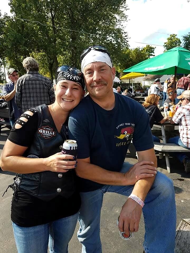 Smiling couple posing at a sunny outdoor festival; woman holds a beer can, both wear bandanas and biker-style clothing.