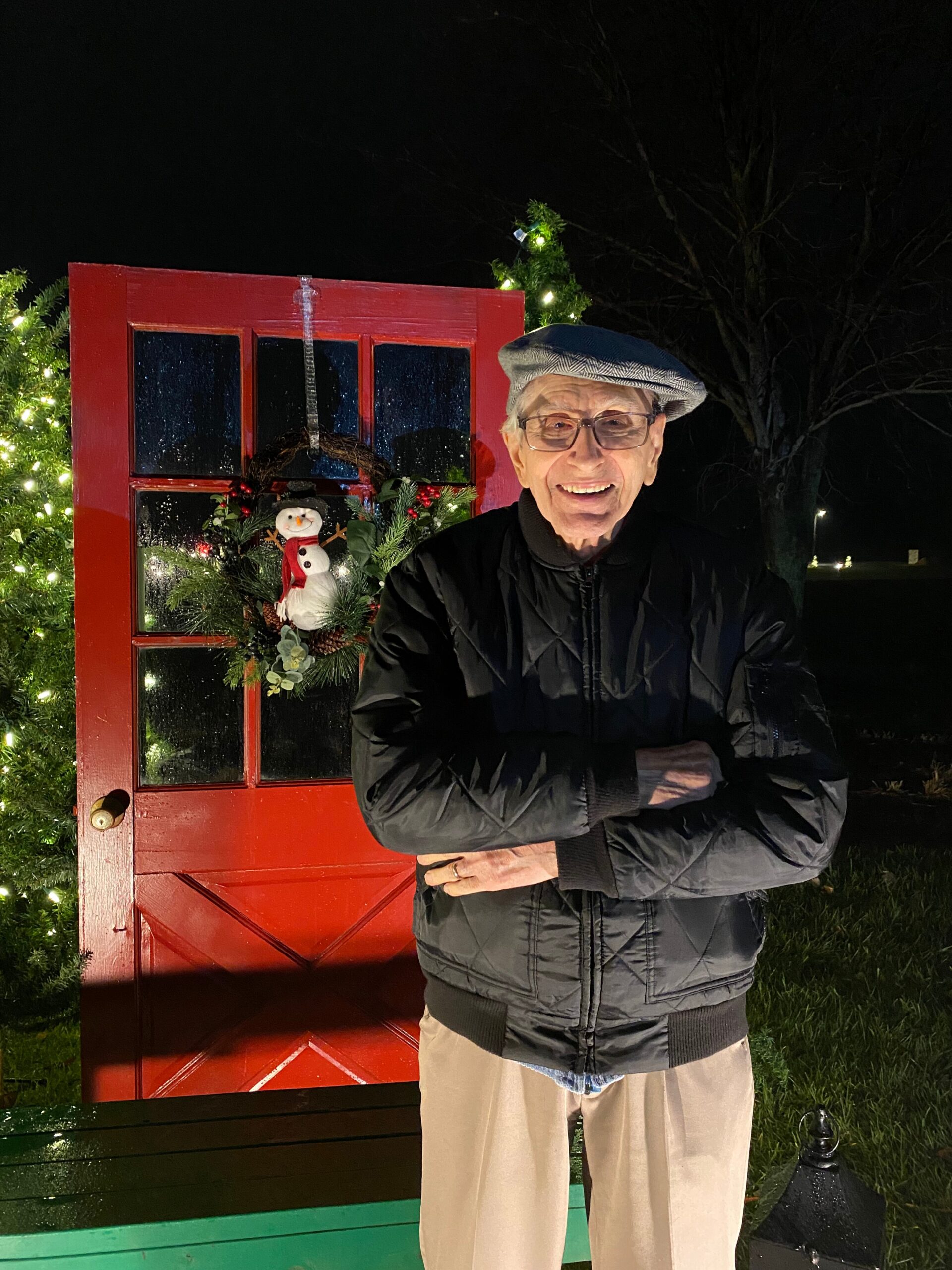 Smiling senior man in a quilted black jacket and beret stands beside a decorated red door with a wreath and Christmas lights at night.