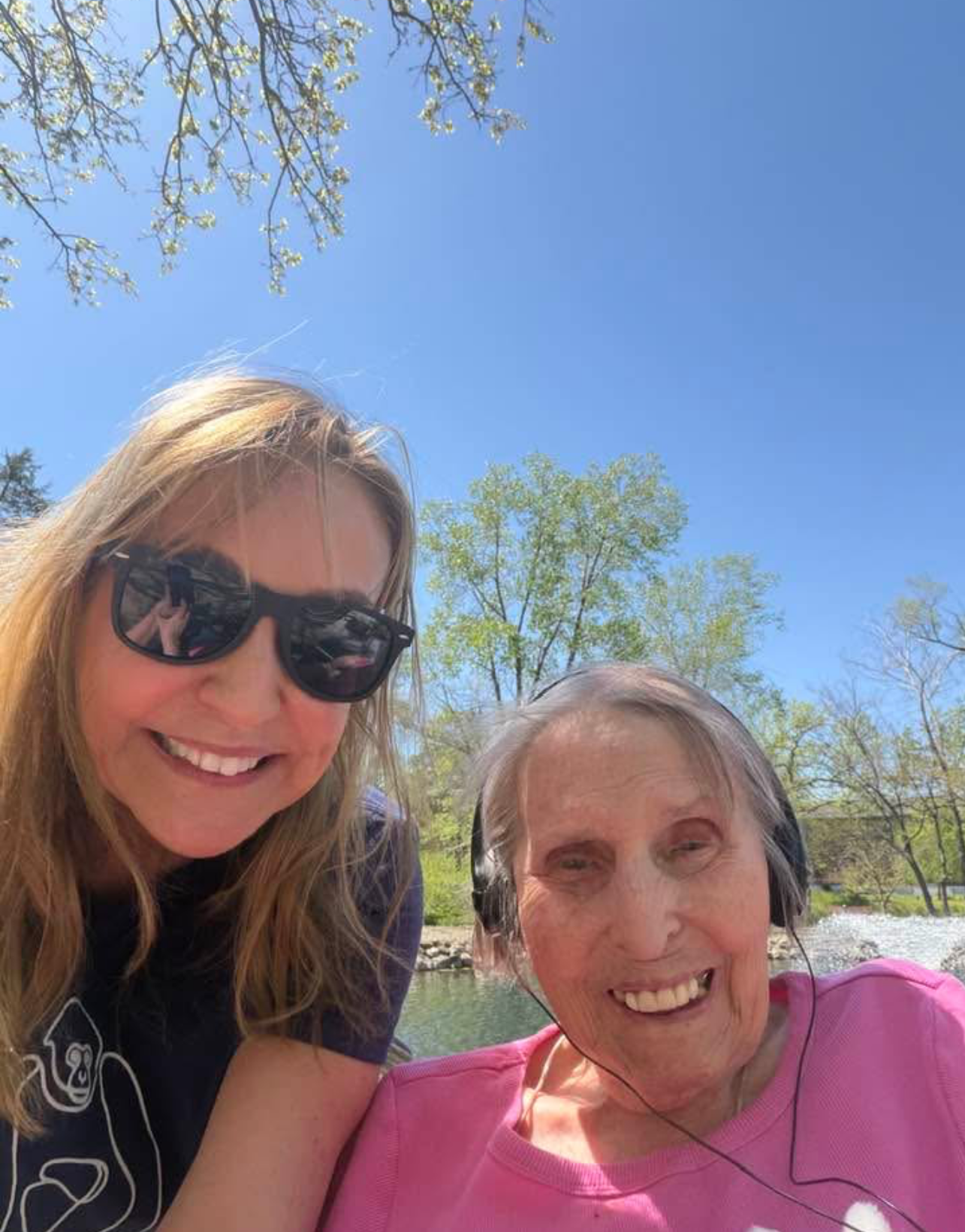Two women smiles for a selfie outdoors on a sunny day, one wearing sunglasses and the other with headphones by trees and blue sky.