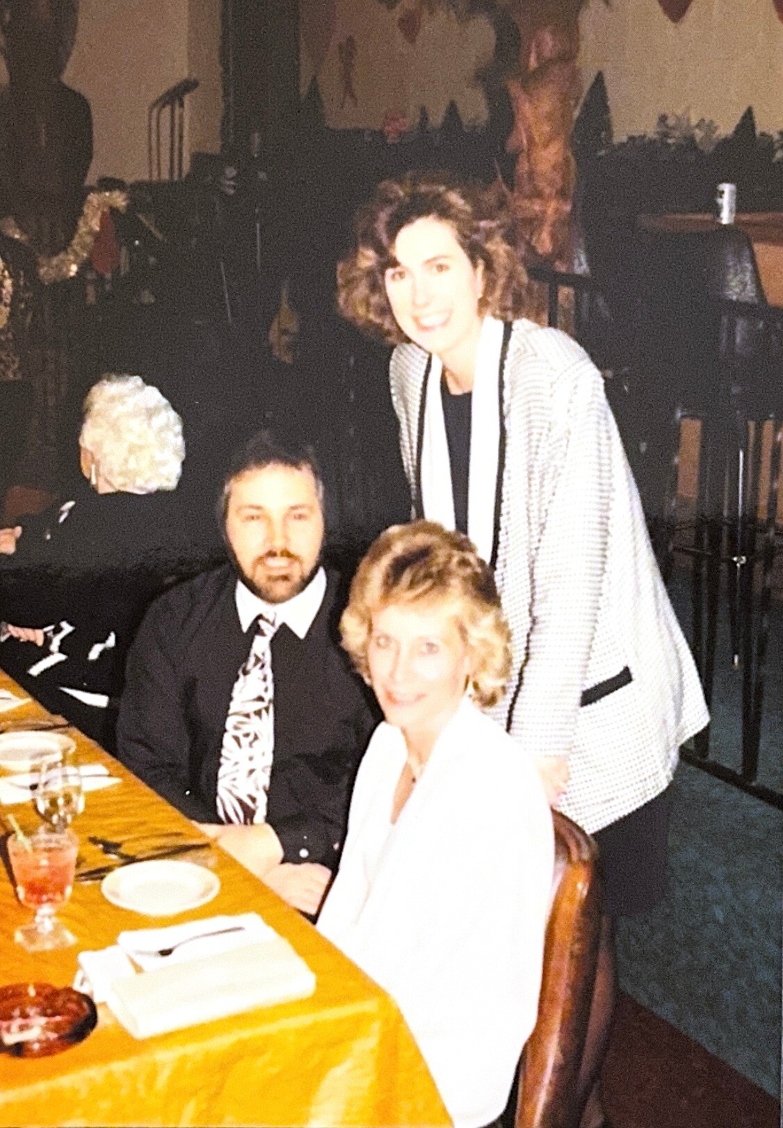 Three adults posing at a dining table: a man in a dark suit, a woman in white, and a woman in a checkered jacket standing behind them, smiling.