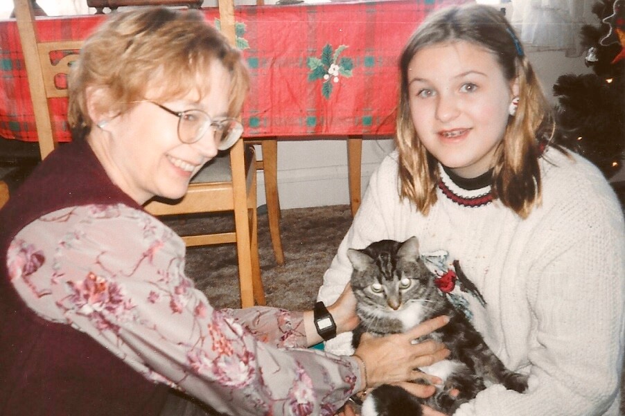 Older woman with glasses and a young girl smiling while holding a cat indoors during a holiday scene.