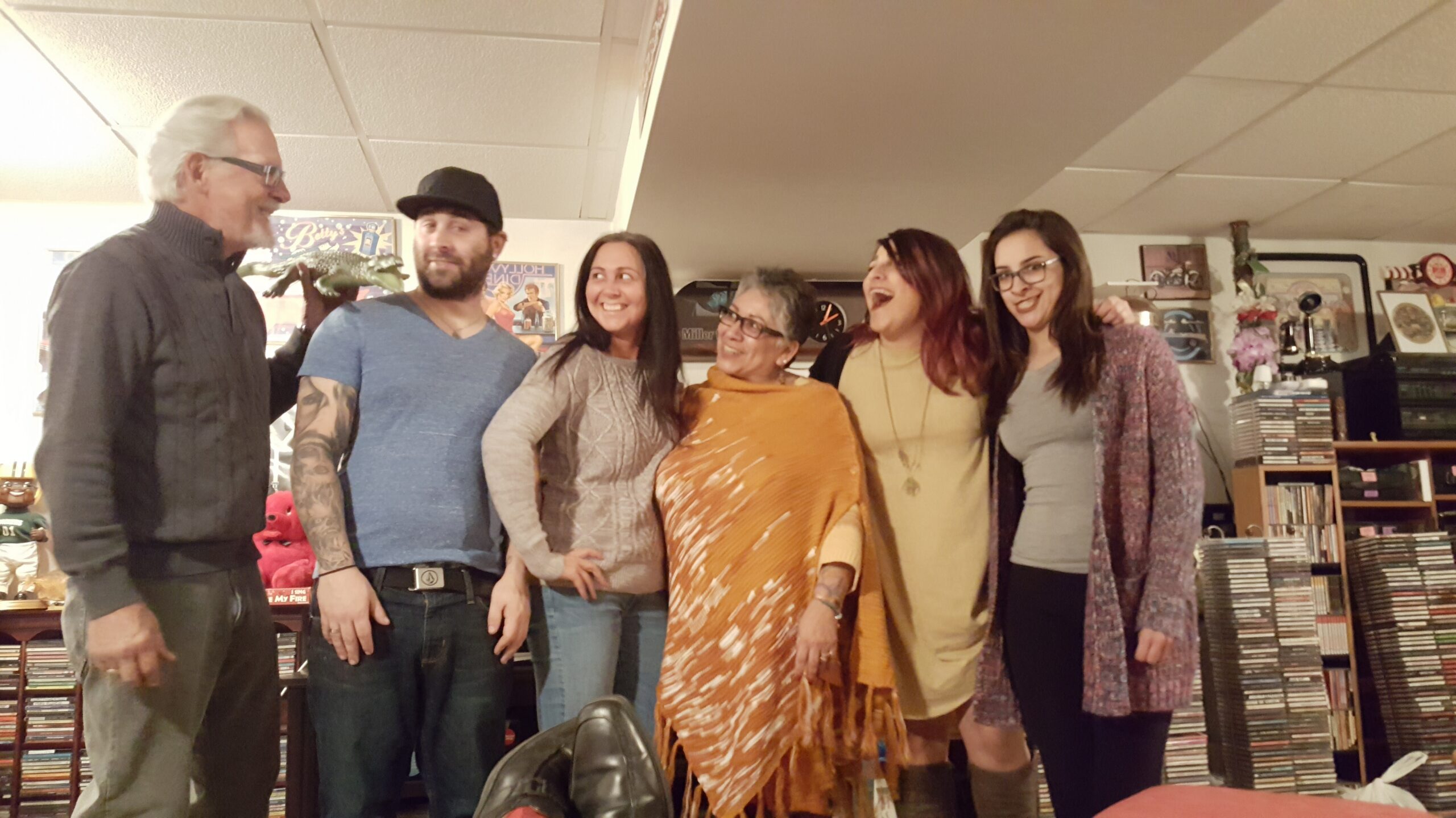 Six adults stand in a row indoors, smiling and posing for a casual group photo with posters and shelves in the background.