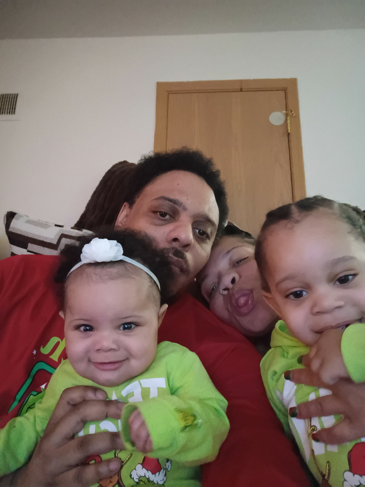 Close-up family selfie: a man with two babies and a young child wearing neon-green outfits indoors, posing for the camera.