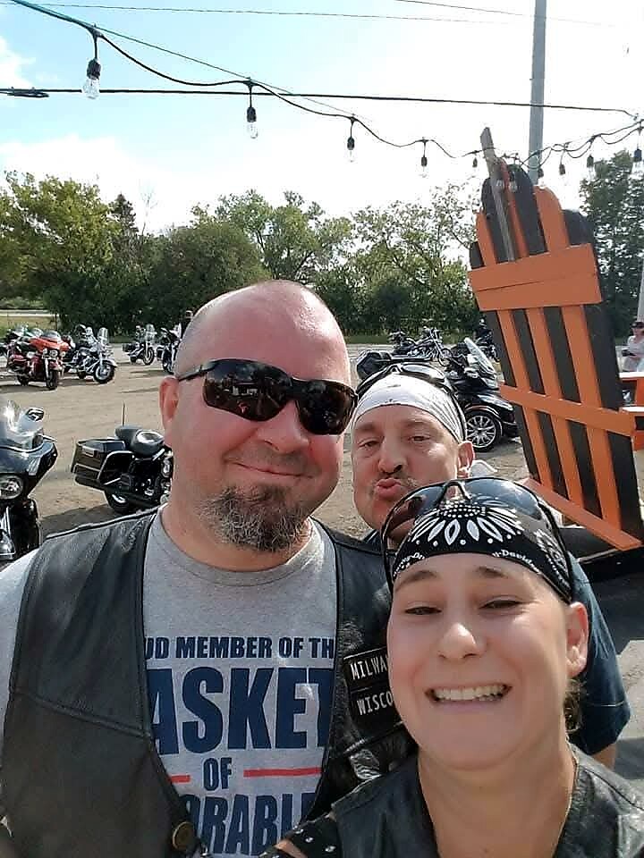Three friends smiling for a selfie at a motorcycle gathering, with motorcycles and string lights in the background.