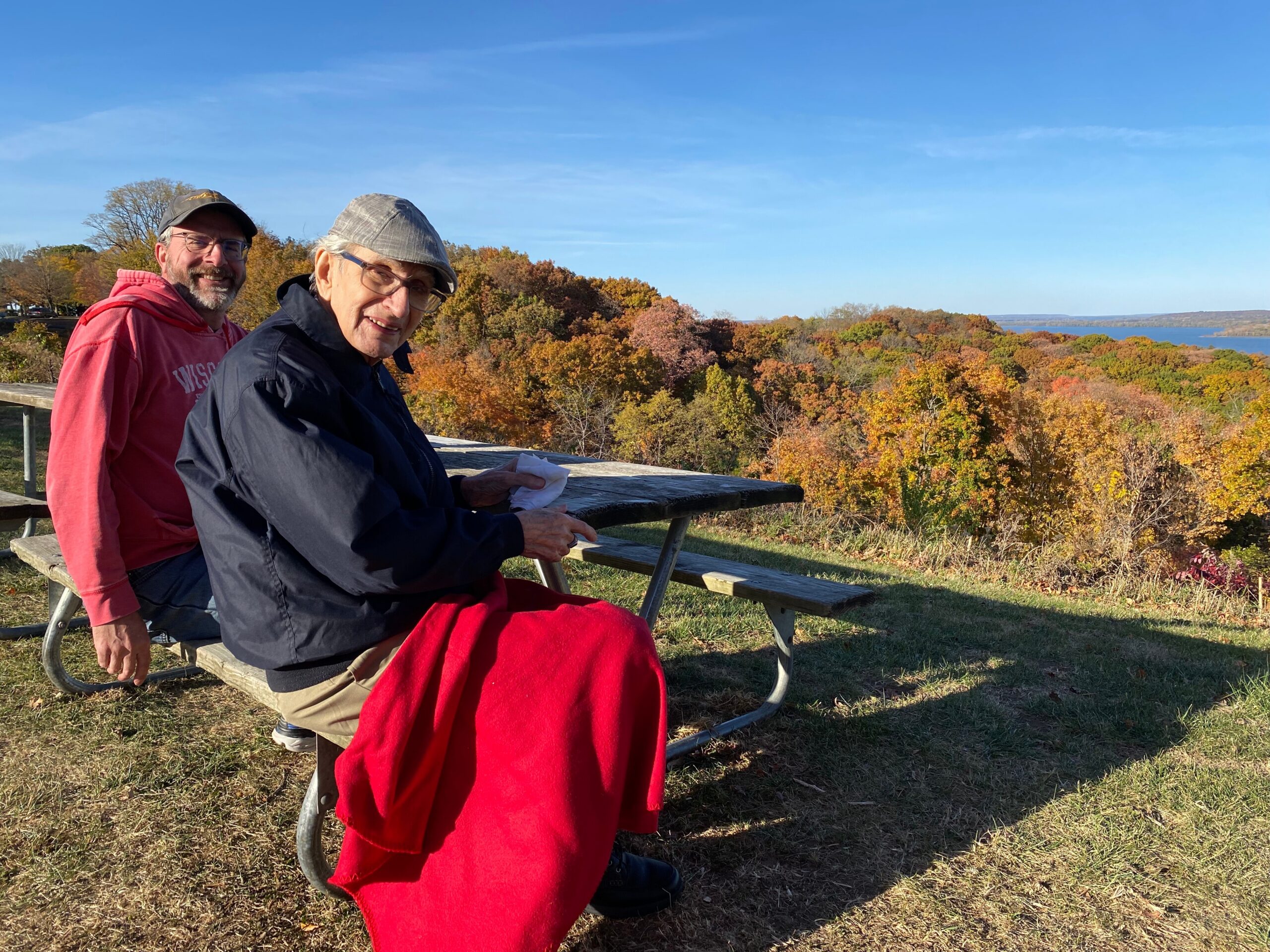 Two men sit on a wooden picnic table overlooking a colorful autumn forest and blue sky; one is old wearing a gray cap, the other in a pink hoodie behind him, smiling at the camera.