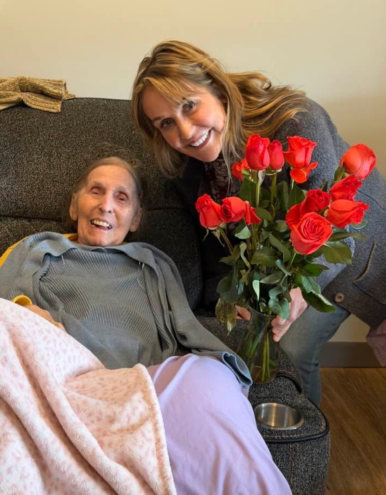Smiling elderly woman resting on a chair with a blanket as a visitor offers a bouquet of red roses nearby.