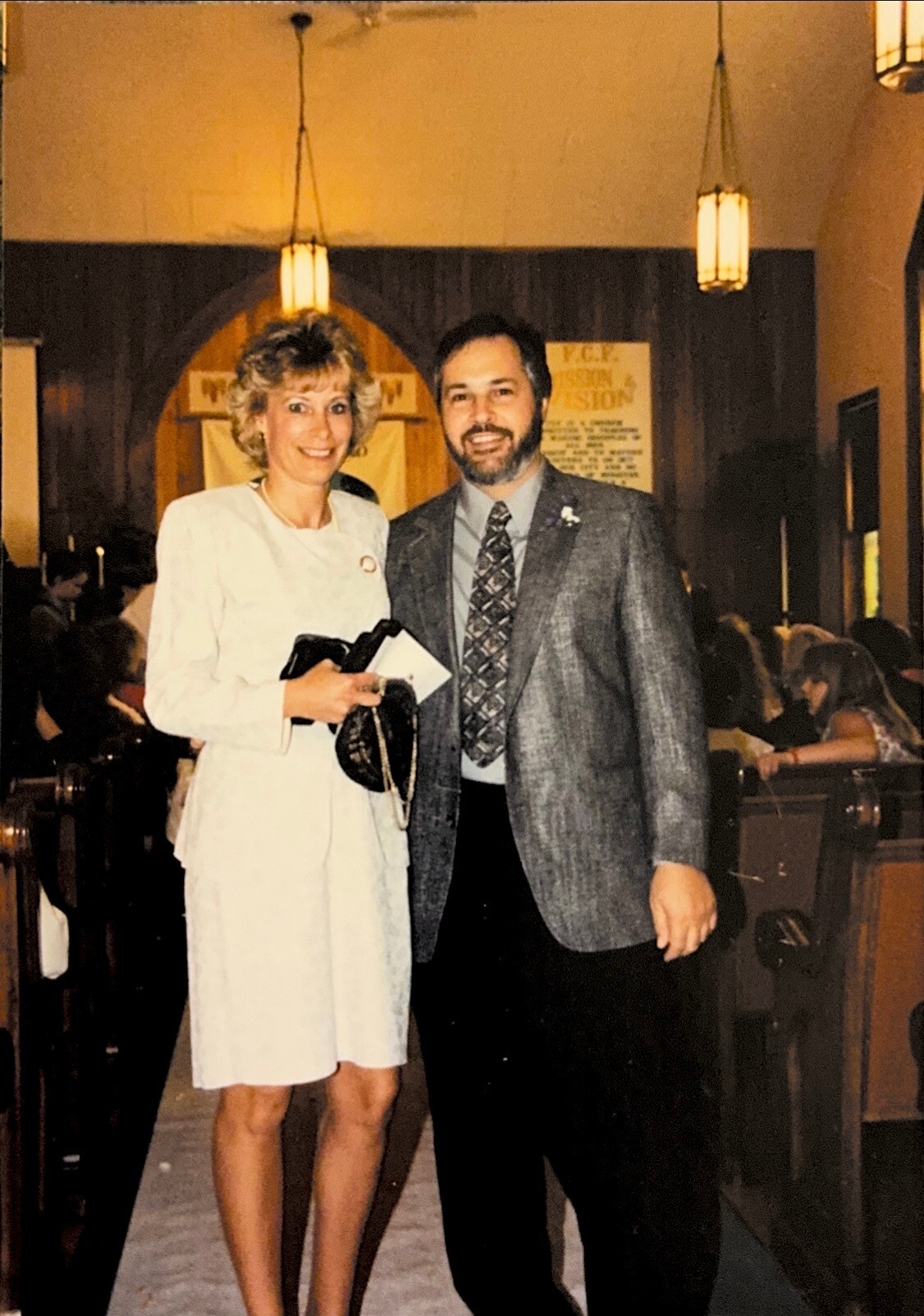 A smiling couple stands together at a formal event, woman in a white dress and man in a gray suit, posing hand in arm in a church-like setting with guests in the background.