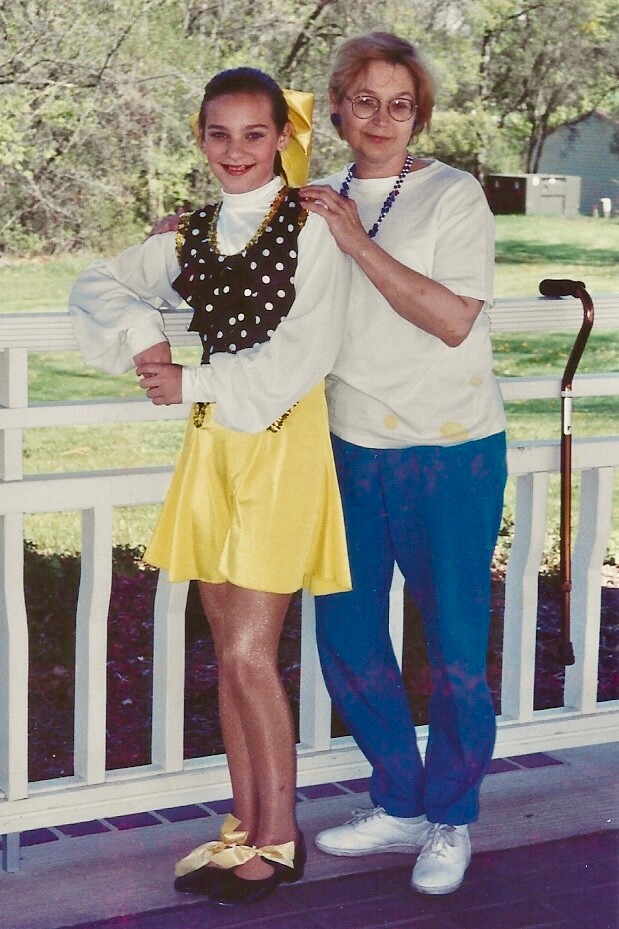 Young girl in a yellow dance dress with a polka-dot bodice and bow poses with an older woman on a porch; a cane rests nearby.