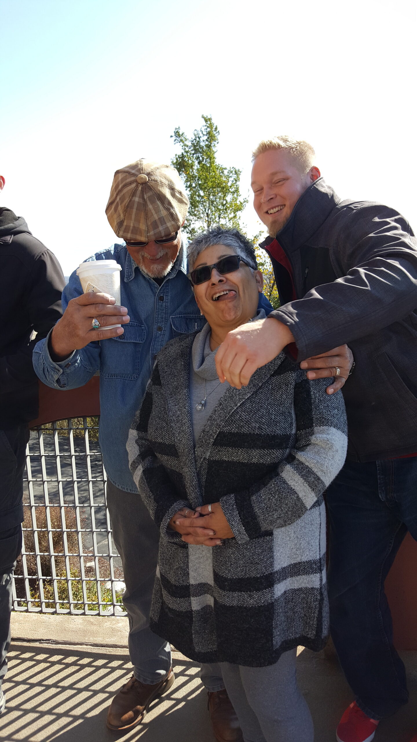 Three adults posing outdoors on a sunny day, smiling; a man holds a coffee cup while two friends hug and pose for the selfie-like photo, wearing casual jackets and sunglasses.