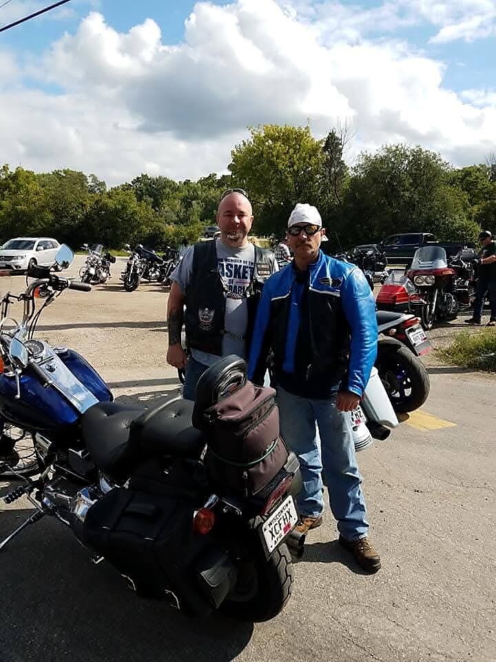 Two men stand beside a large touring motorcycle with saddlebags in a parking lot, other bikes and trees in the background.