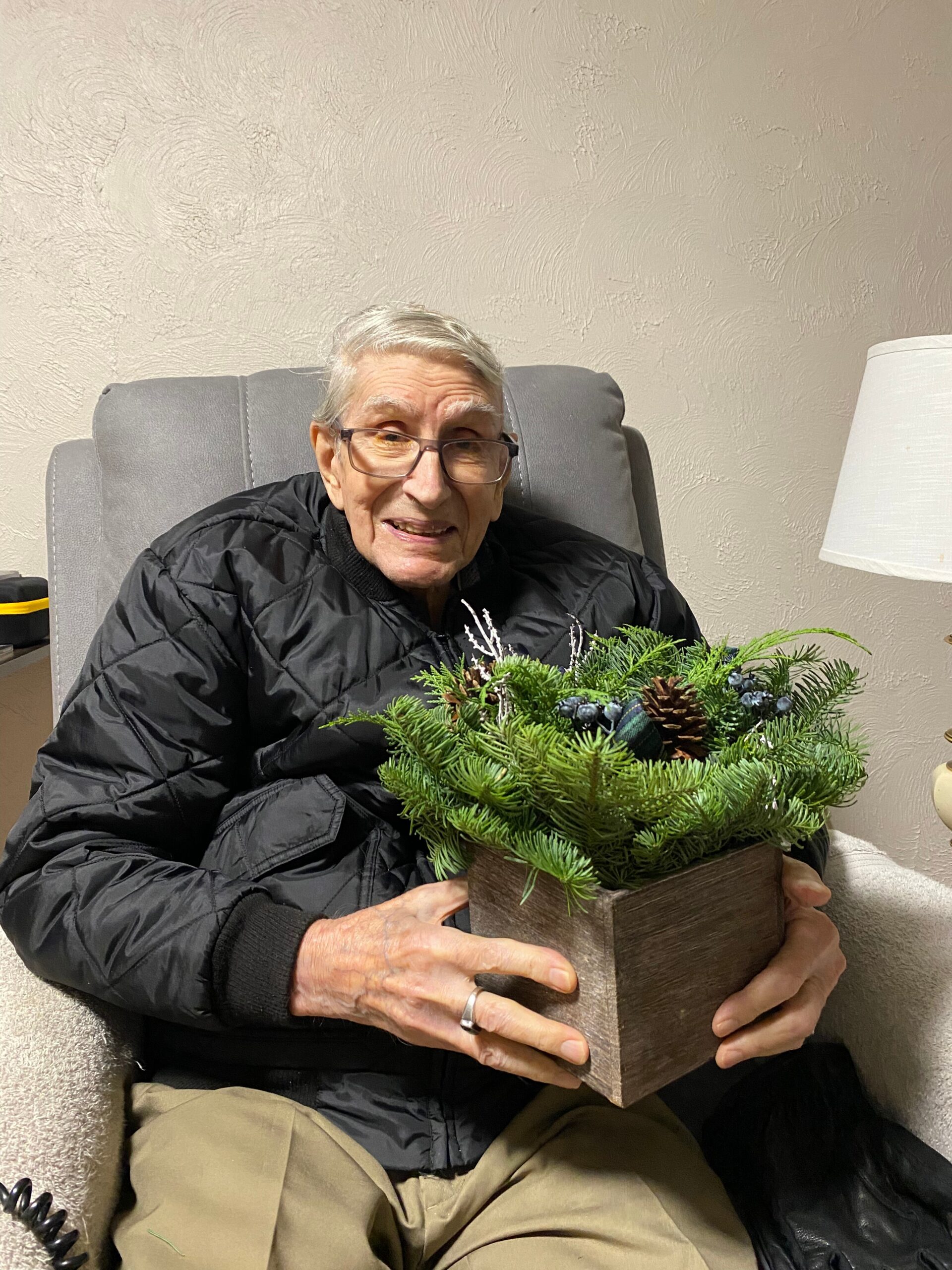 Older man with gray hair and glasses sits in a gray armchair, holding a wooden box with a festive evergreen arrangement (pine branches, pinecones).