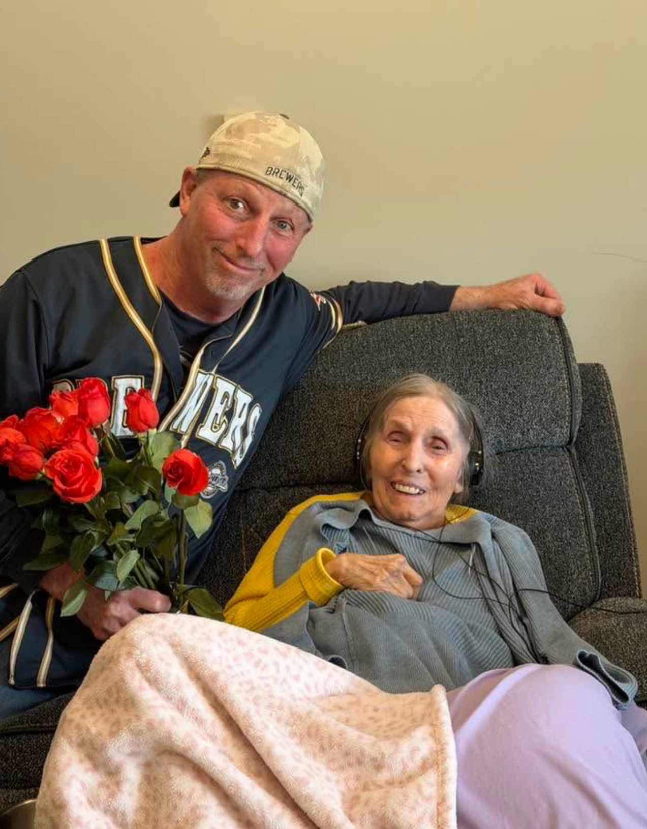 Man in a baseball jersey hands a bouquet of red roses to an elderly woman seated on a couch, both smiling at the camera.