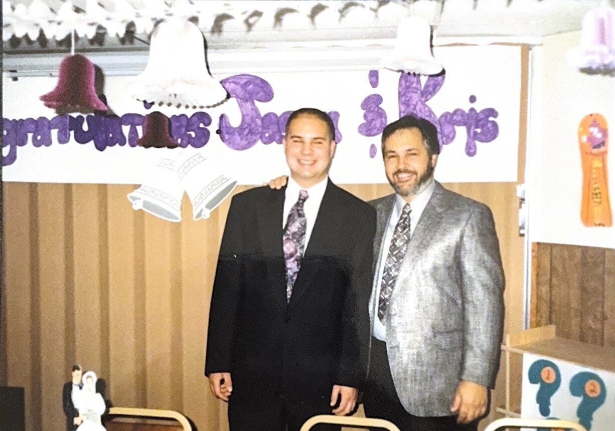Two men in formal attire posing together at a graduation ceremony with a purple banner behind them.
