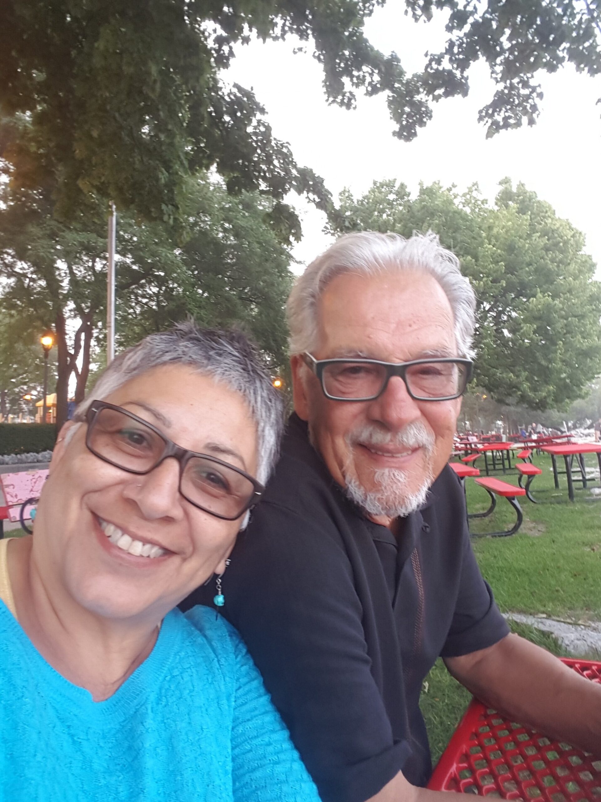 Smiling couple taking a selfie in a park with green trees and red picnic tables behind them to the right.