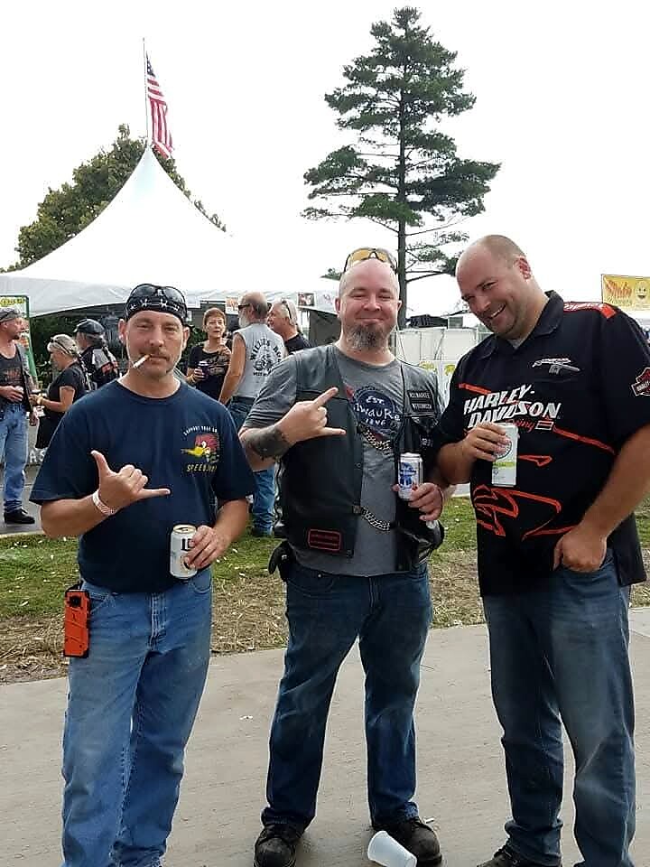 Three men at an outdoor festival posing for the camera; one with a cigarette, two showing rock-on hand signs and holding drinks, a tent and trees behind them.