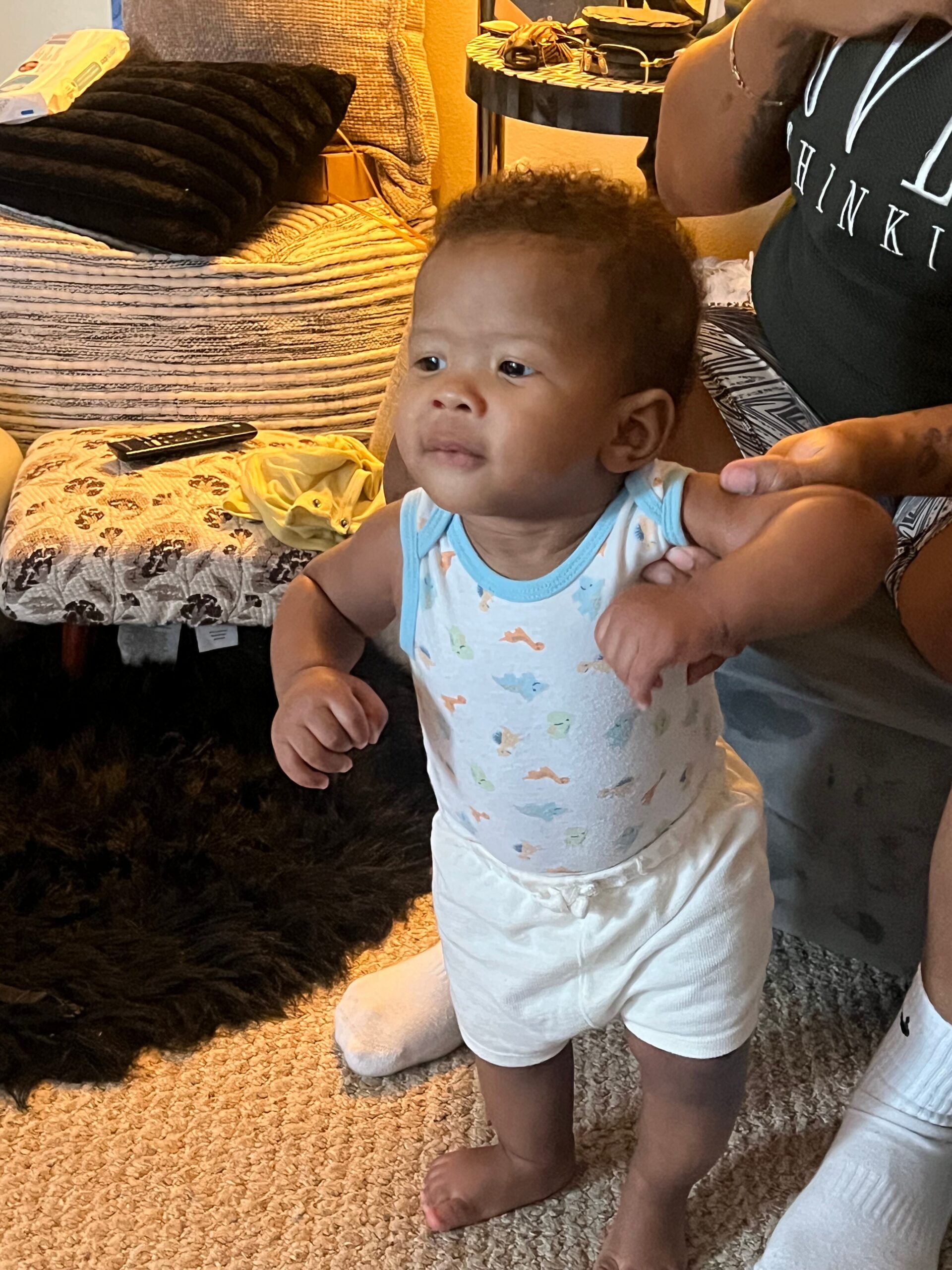 Young child standing with assistance indoors, wearing a light blue dinosaur-print onesie and white shorts.