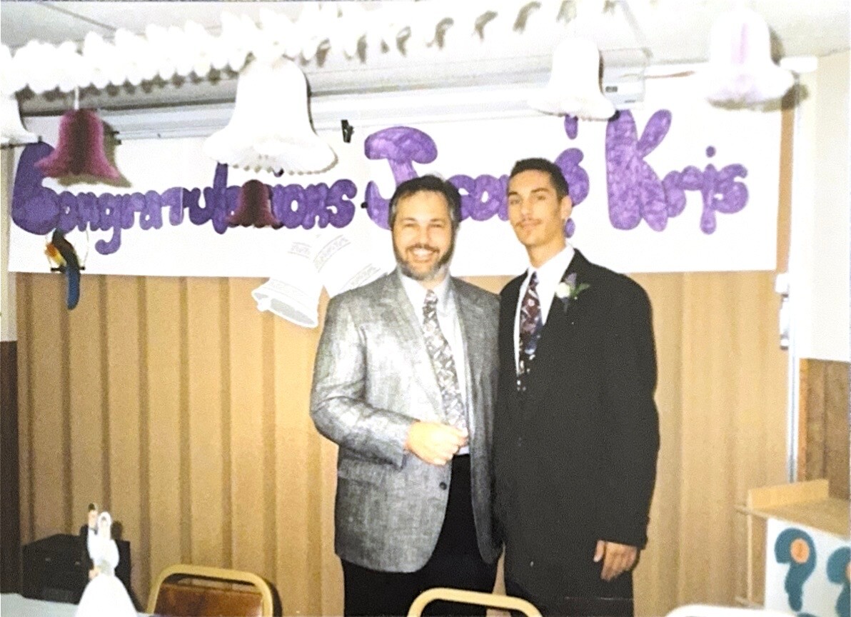 Two men in suits posing together at a celebration, with a purple 'Congratulations' banner in the background.