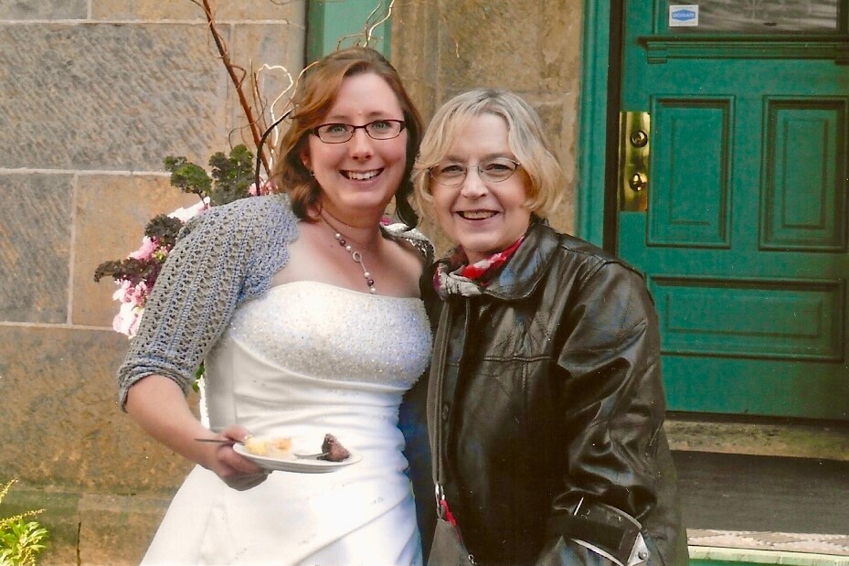 Bride in a white gown with a grey shawl, holding a plate with cake, posing with a guest outside a stone building with a green door.