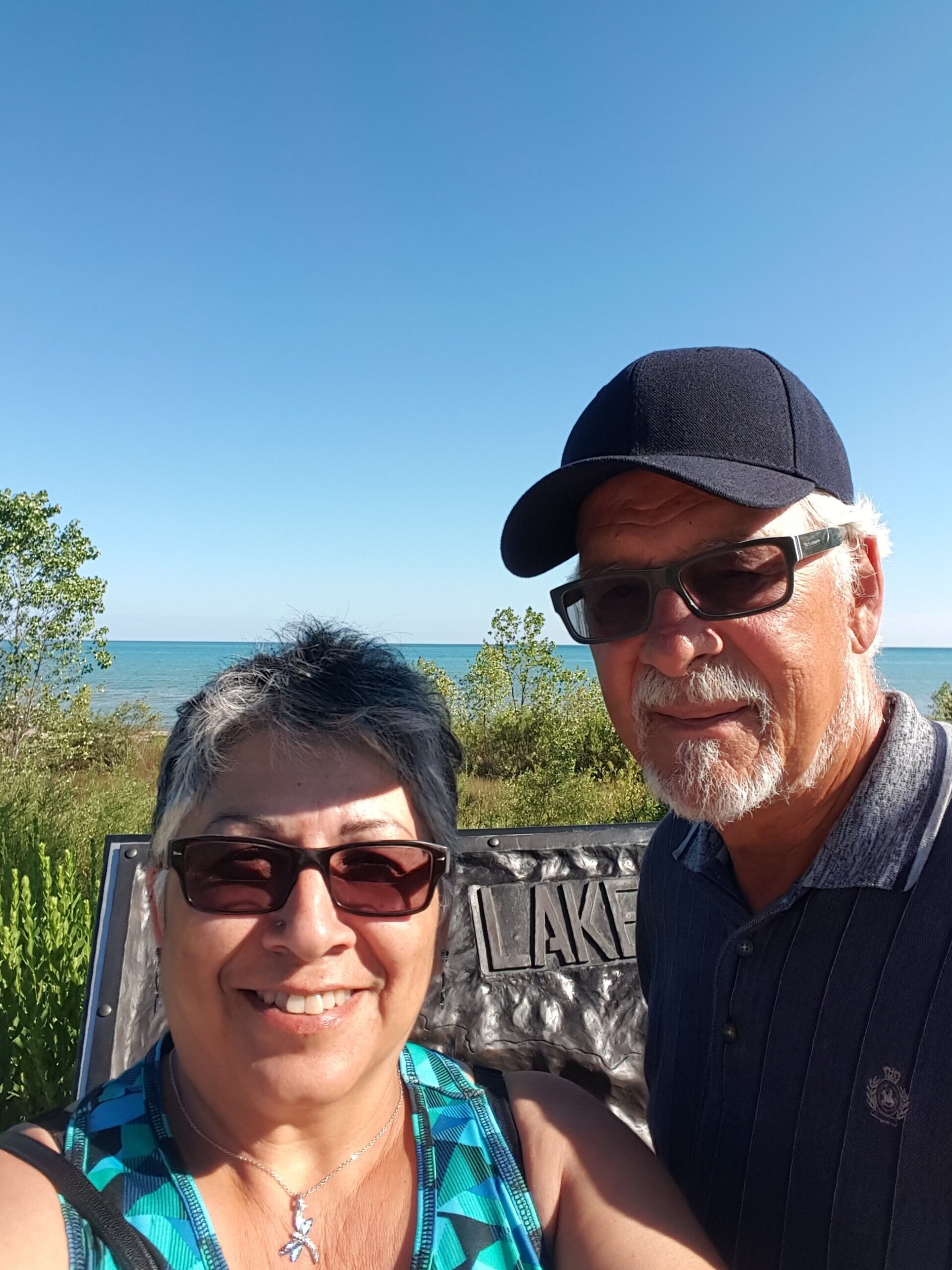 Smiling couple taking a selfie outdoors by a lake under a clear blue sky; man wearing a black cap and sunglasses, woman in a teal patterned top.