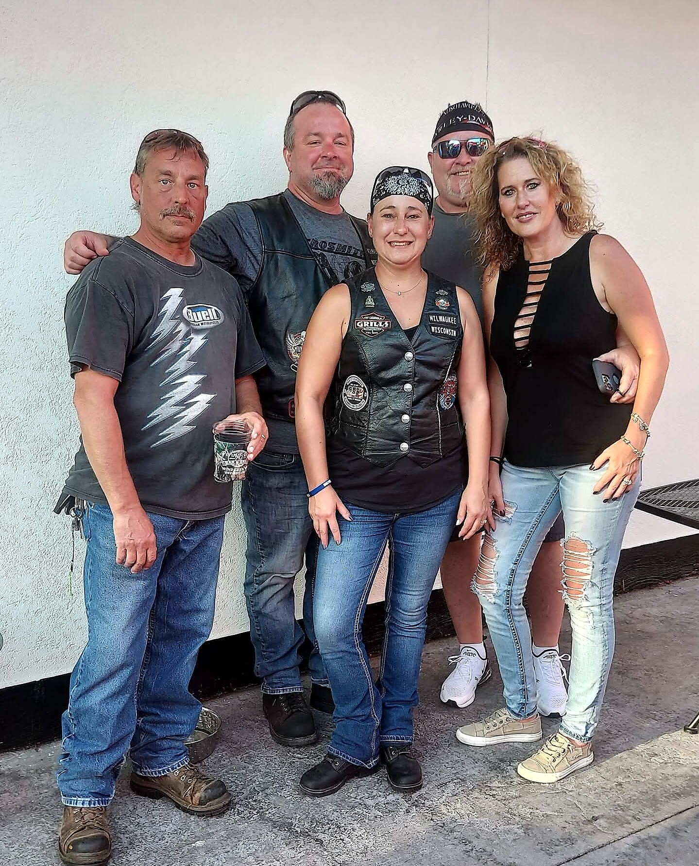 Five adults pose together against a light wall, two women in front and three men in casual biker-style clothing, smiling at the camera.
