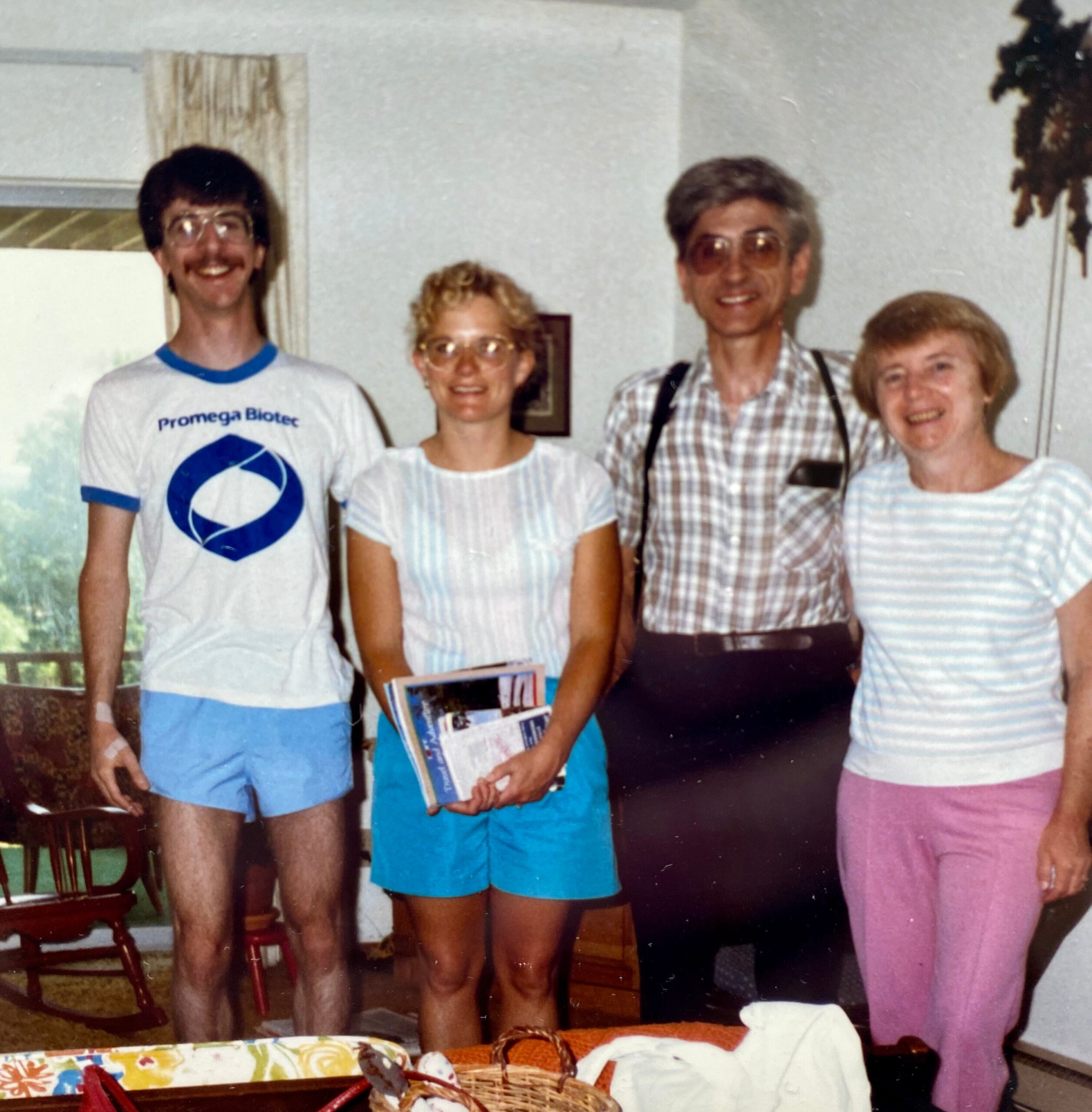 Four adults pose together in a living room, smiling at the camera in casual 1980s outfits.