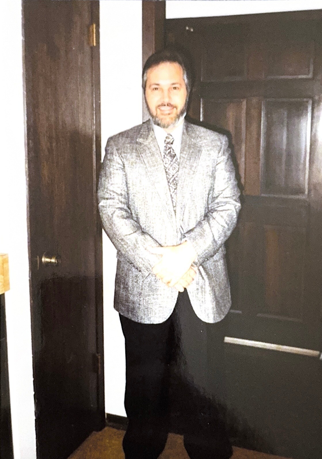 Man in a gray blazer and patterned tie stands in a doorway, hands clasped in front, smiling at the camera.