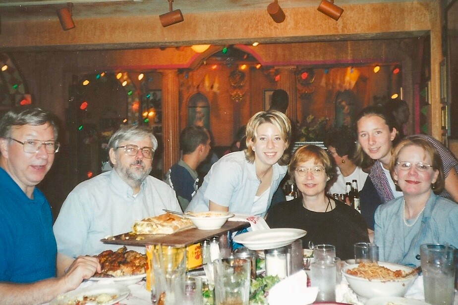 Group of six adults seated around a festive restaurant table with plates of food and drinks, smiling at the camera.