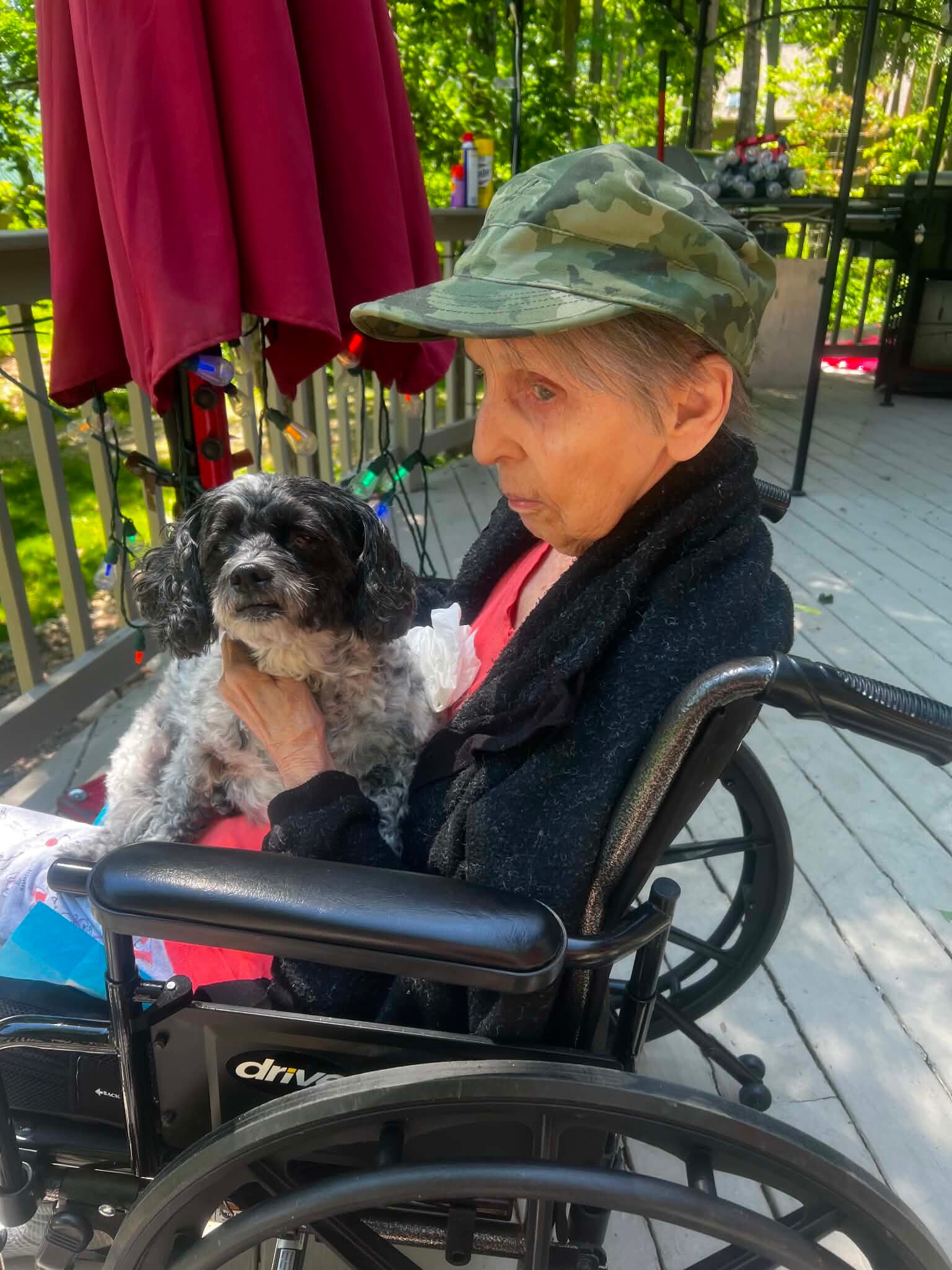 Older person in a wheelchair holding a small dog outdoors on a deck.
